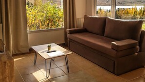 Una sala de estar con un sofá marrón, una mesita y vistas a un lago y montañas a través de amplios ventanales.
