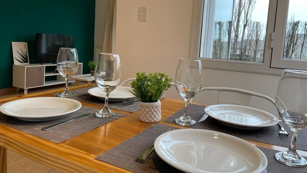 Una mesa de comedor dispuesta con platos, vasos y cubiertos, una pequeña planta y un televisor al fondo.