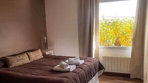 Un dormitorio con una cama grande, una mesita de noche, una lámpara colgante, una ventana con cortinas y vistas al exterior.