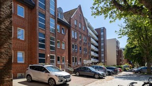 A multi-story residential building with brick facades, a parking area with several cars, and a tree providing shade.