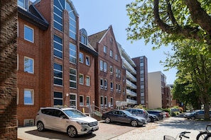 Un immeuble résidentiel à plusieurs étages, doté de façades en briques, un espace de stationnement avec plusieurs voitures et un arbre qui offre de l’ombre.