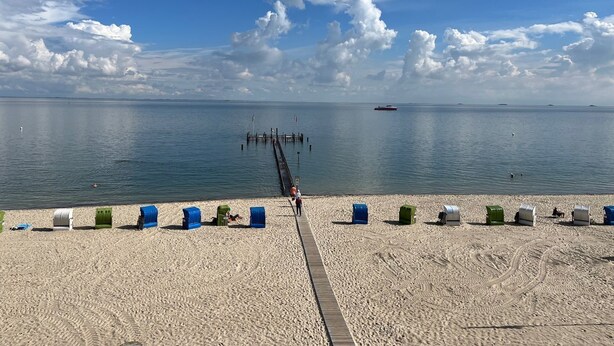 A beach with a pier extending into calm waters, a distant boat, and beach chairs lined up along the shore.