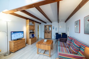 A living room with a wooden TV stand, a dining table, a colorful sofa, and a kitchen area with wooden cabinets.