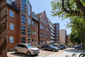 A multi-story residential building with brick facades, a parking area with several cars, and a tree providing shade.