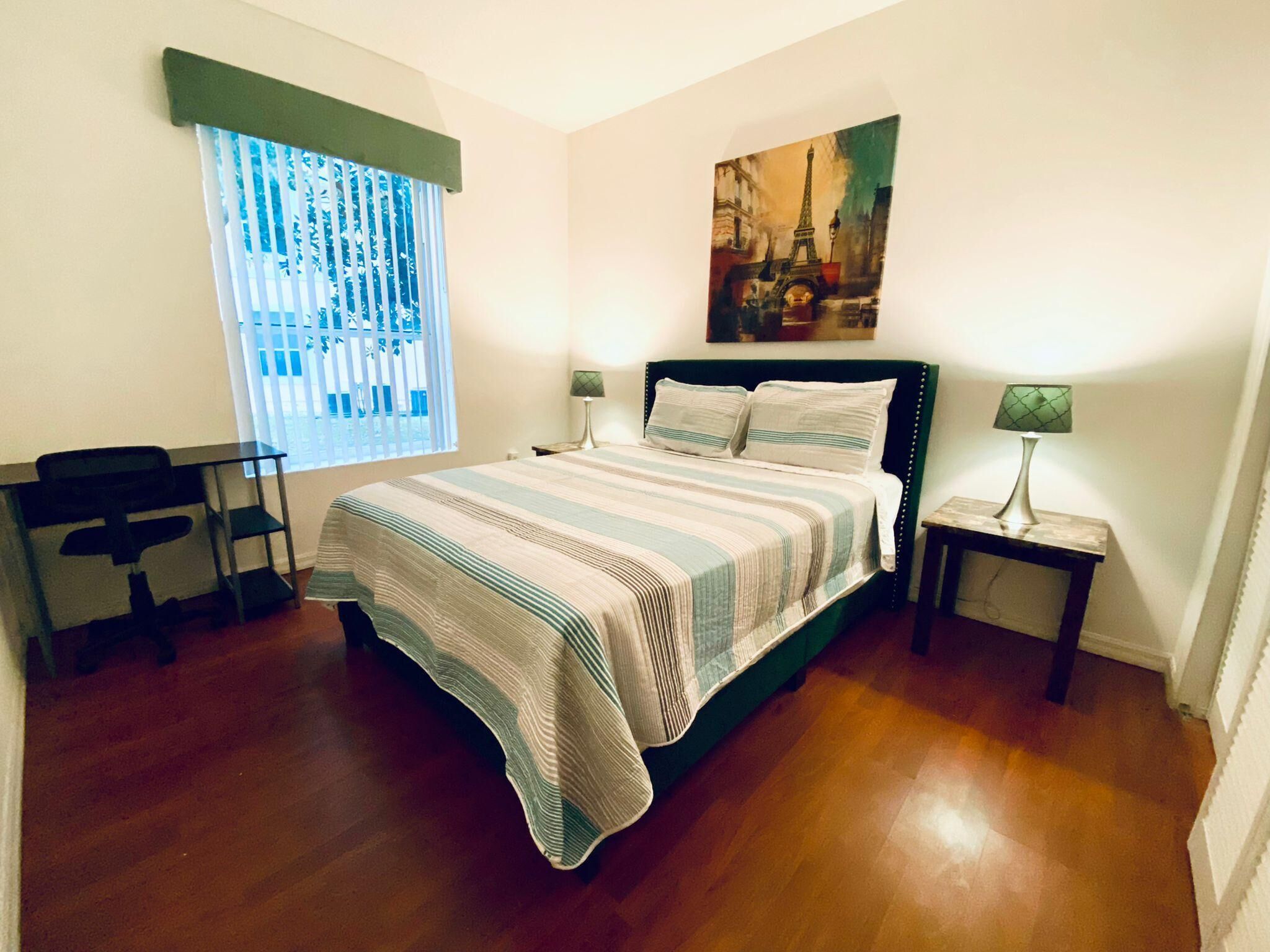 A bedroom with a bed, a desk, a chair, a lamp, and a painting of the Eiffel Tower.