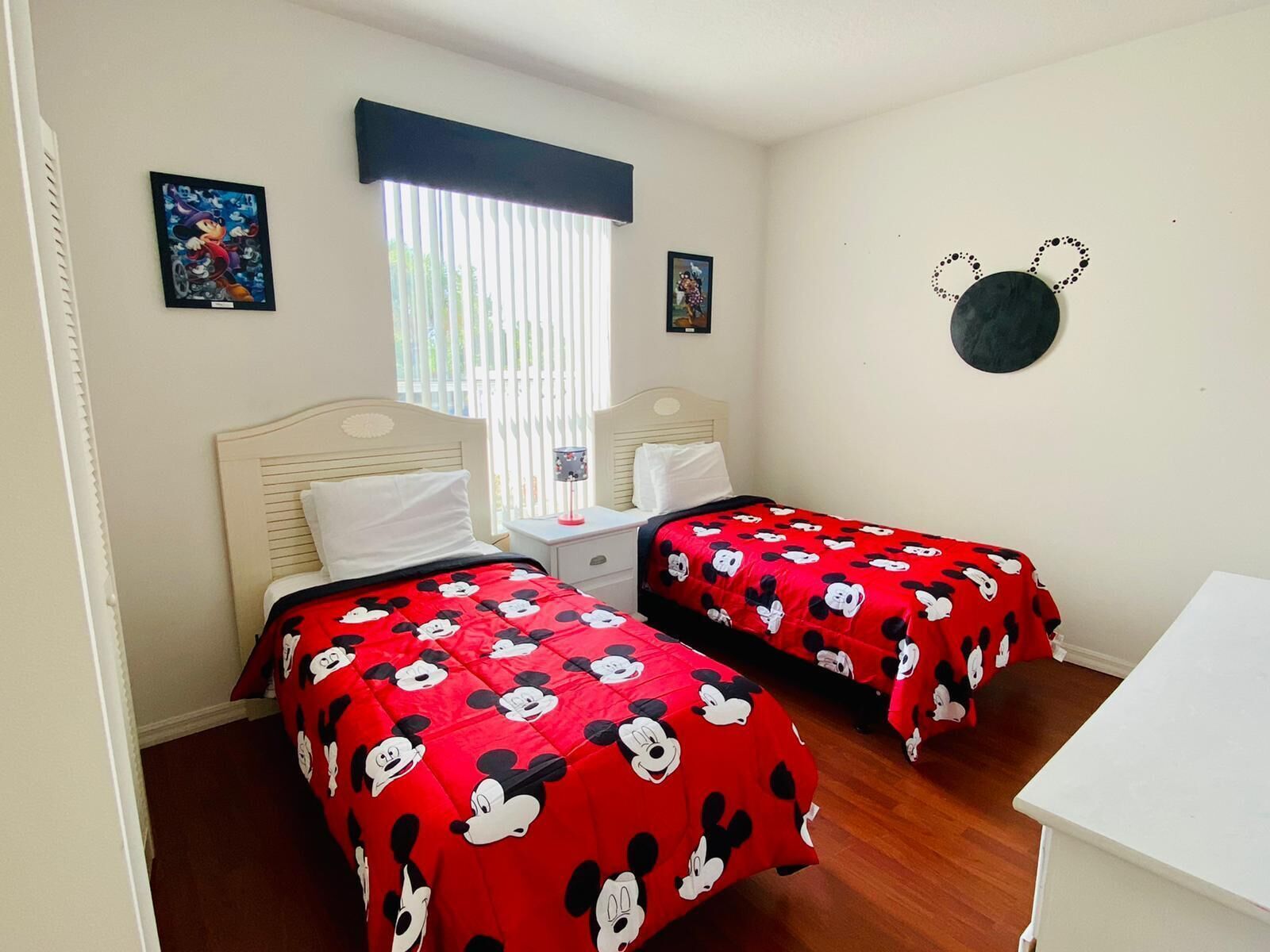 Una habitación con dos camas, cada una con ropa de cama roja con estampado de Mickey Mouse, una ventana con persianas y un reloj de pared.