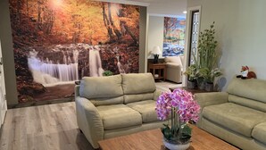A living room with a beige sofa, a wooden coffee table with pink orchids, and a large wall mural of a waterfall.