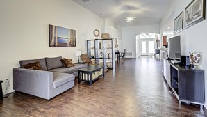A spacious living room with a grey sofa, a coffee table, and a TV stand. There is a bookshelf and a dining area in the background. - Gated Community, Private Pool and Game Room Near Disney!!! (Kissimmee)