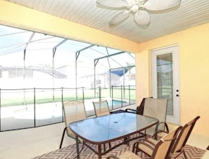 Um alpendre coberto com mesa e cadeiras, um ventilador de teto e vista para uma piscina e casas.