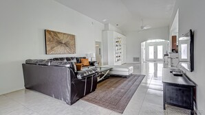 A modern living room with a black leather sofa, a coffee table, and a large abstract painting. There is a white sofa in the background, a bookshelf, and a kitchen area visible through an archway. - Gated Community, Private Pool and Game Room Near Disney!!! (Kissimmee)