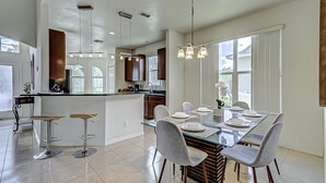 A modern kitchen with a central island, bar stools, and a dining area with a glass table and chairs. - Gated Community, Private Pool and Game Room Near Disney!!! (Kissimmee)