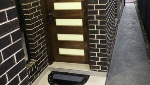 Una casa moderna con fachada de ladrillo oscuro, una puerta de madera con paneles de vidrio horizontales y un sendero de concreto que lleva a un felpudo negro.