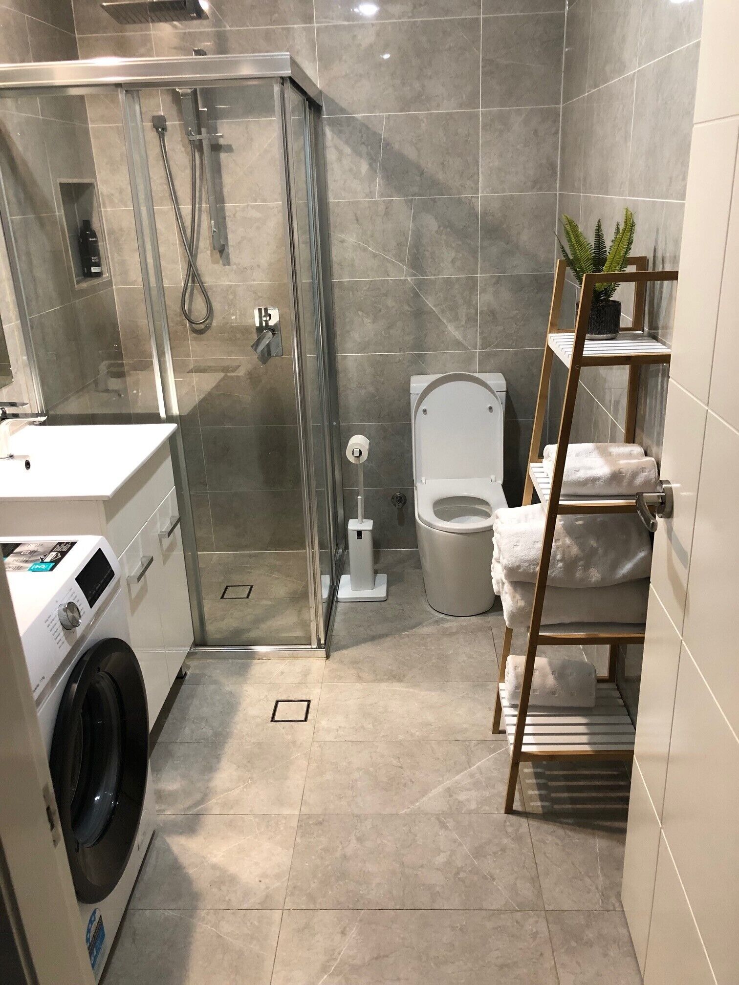Et moderne badeværelse med en glasafskærmet brusekabine, toilet, vaskemaskine og et håndklædestativ med håndklæder.