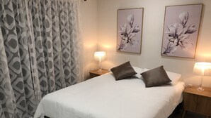 Una cama bien tendida con ropa blanca y dos almohadas marrones, flanqueada por dos mesitas de noche con lámparas, en una habitación con cortinas estampadas y dos cuadros florales colgados en la pared.
