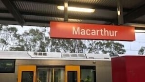 Un andén de estación de tren con un letrero rojo que dice 'Macarthur'.