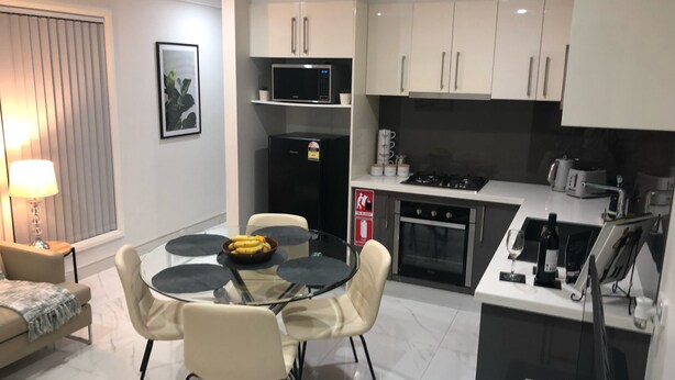 Una cocina moderna con armarios blancos, un refrigerador negro y una estufa. Hay una mesa de comedor con sillas y un bol de fruta.