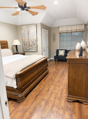 A bedroom with a bed, a desk, a chair, a ceiling fan, a painting, and a window with blinds.
