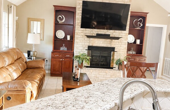 A living room with a leather sofa, a fireplace, a TV mounted above it, and a kitchen area with a marble countertop and sink.