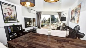 A modern living room with a wooden dining table, white sofas, a TV, and a large window with a view of the outdoors. - Stunning 1 bed apt Benalmadena (Benalmádena)