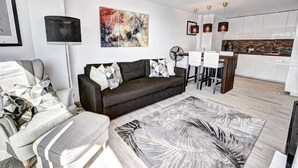 A modern living room with a sofa, a coffee table, and a large abstract painting. There is a kitchen area with white cabinets and a wooden countertop. - Stunning 1 bed apt Benalmadena (Benalmádena)