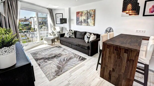 A modern living room with a dark grey sofa, a wooden dining table, and a large window offering a view of the city. - Stunning 1 bed apt Benalmadena (Benalmádena)
