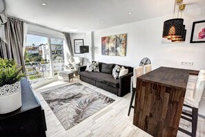 A modern living room with a dark grey sofa, a wooden dining table, and a large window offering a view of the city.