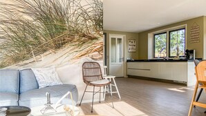 A modern living room with a grey sofa, a white coffee table, and a wicker chair. A kitchen with white cabinets and a large window is visible in the background. - 5 bedroom awesome home in Groote Keeten (Groote Keeten)