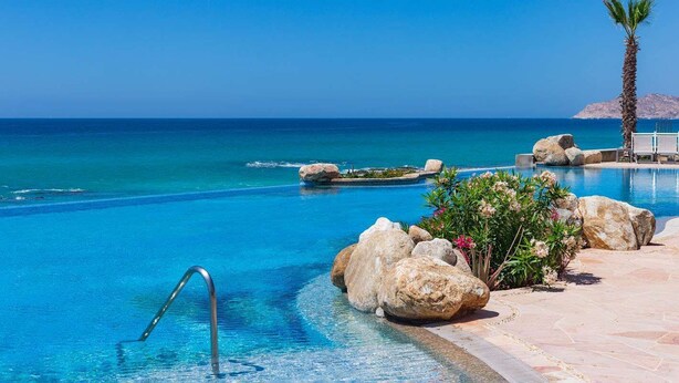Ein klares, blaues Schwimmbecken mit einer Metallleiter, umgeben von großen Felsen und einer Palme, mit Blick auf das Meer und einen entfernten Berg.
