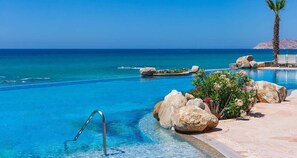 Ein klares, blaues Schwimmbecken mit einer Metallleiter, umgeben von großen Felsen und einer Palme, mit Blick auf das Meer und einen entfernten Berg.