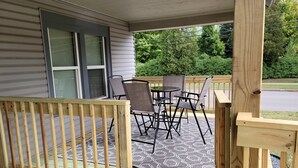 Een houten terras met een tafel, stoelen, een kamerkleed met patroon en uitzicht op groen en een weg.