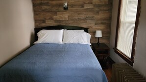 Een bed met een blauwe spreideken, witte kussens, een houten hoofdbord, een nachtkastje met een lamp en een raam met jaloezieën.