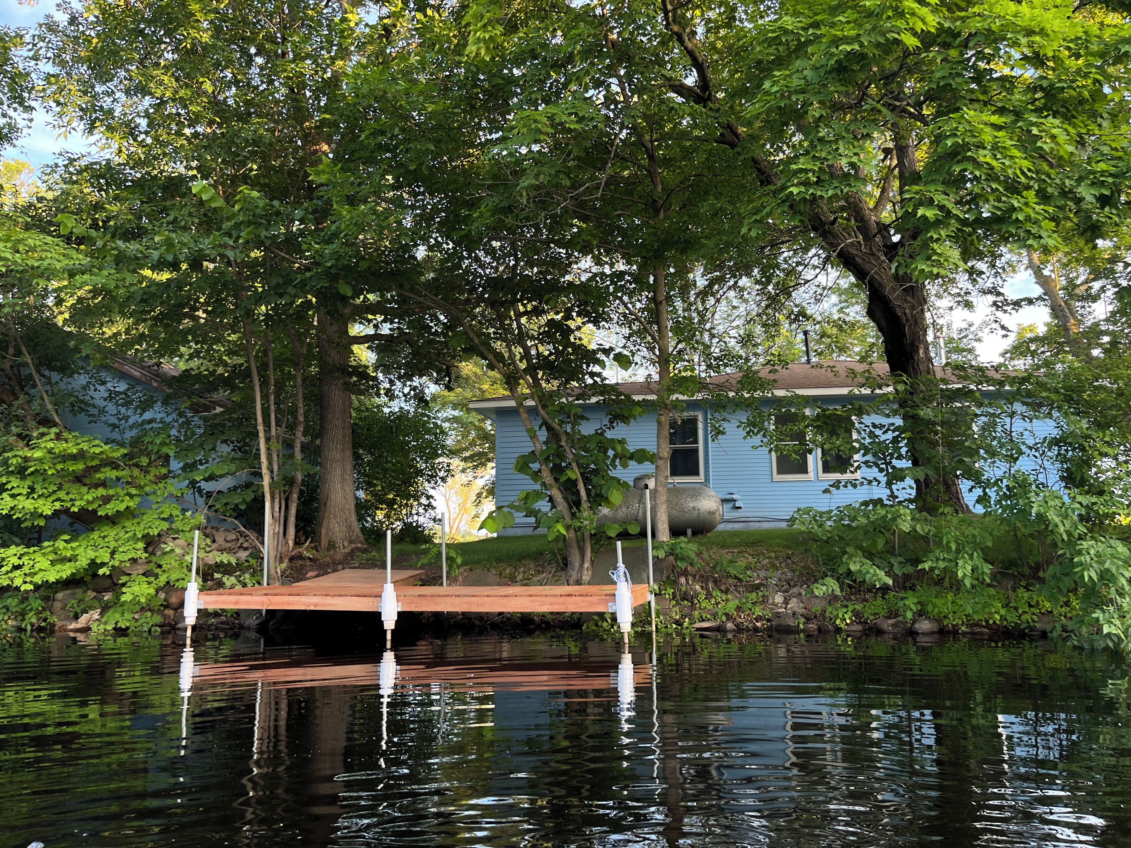 Une jetée en bois s’étendant sur une étendue d’eau calme, entourée d’arbres verdoyants et avec une maison bleue en arrière-plan.