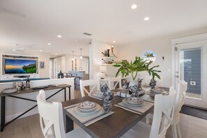 A modern kitchen with a dining area, featuring a large flat-screen TV, white cabinetry, and a central island with bar stools.