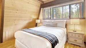 Une chambre avec un plafond en bois, une grande fenêtre, un lit avec une literie blanche, une table de chevet avec une lampe et une commode avec des tiroirs.