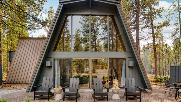 Une structure moderne en bois, de forme triangulaire, dotée de grandes fenêtres en verre, d’un espace extérieur aménagé et surplombant une forêt.