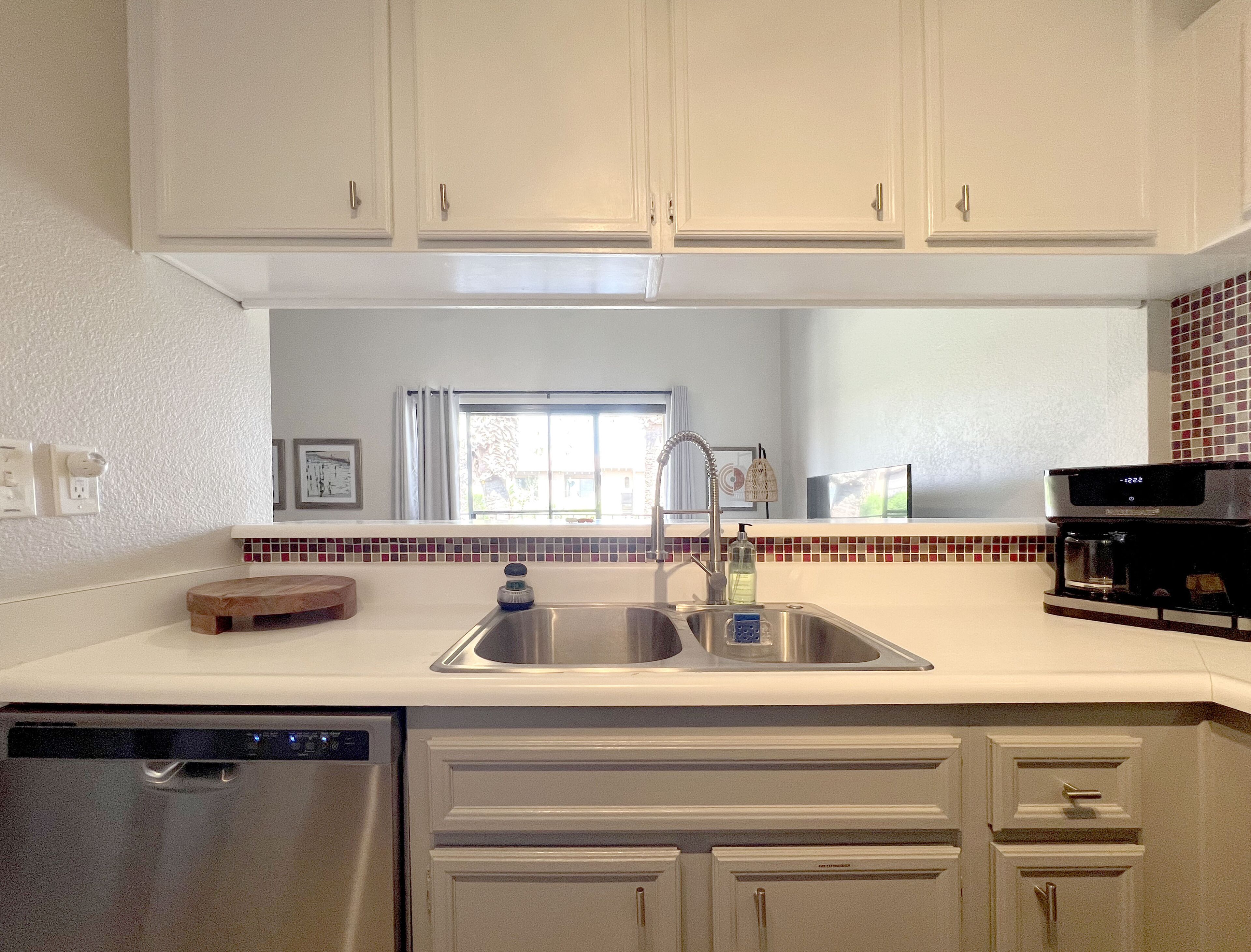 Una cocina con armarios blancos, un lavamanos de acero inoxidable, lavavajillas y cafetera.