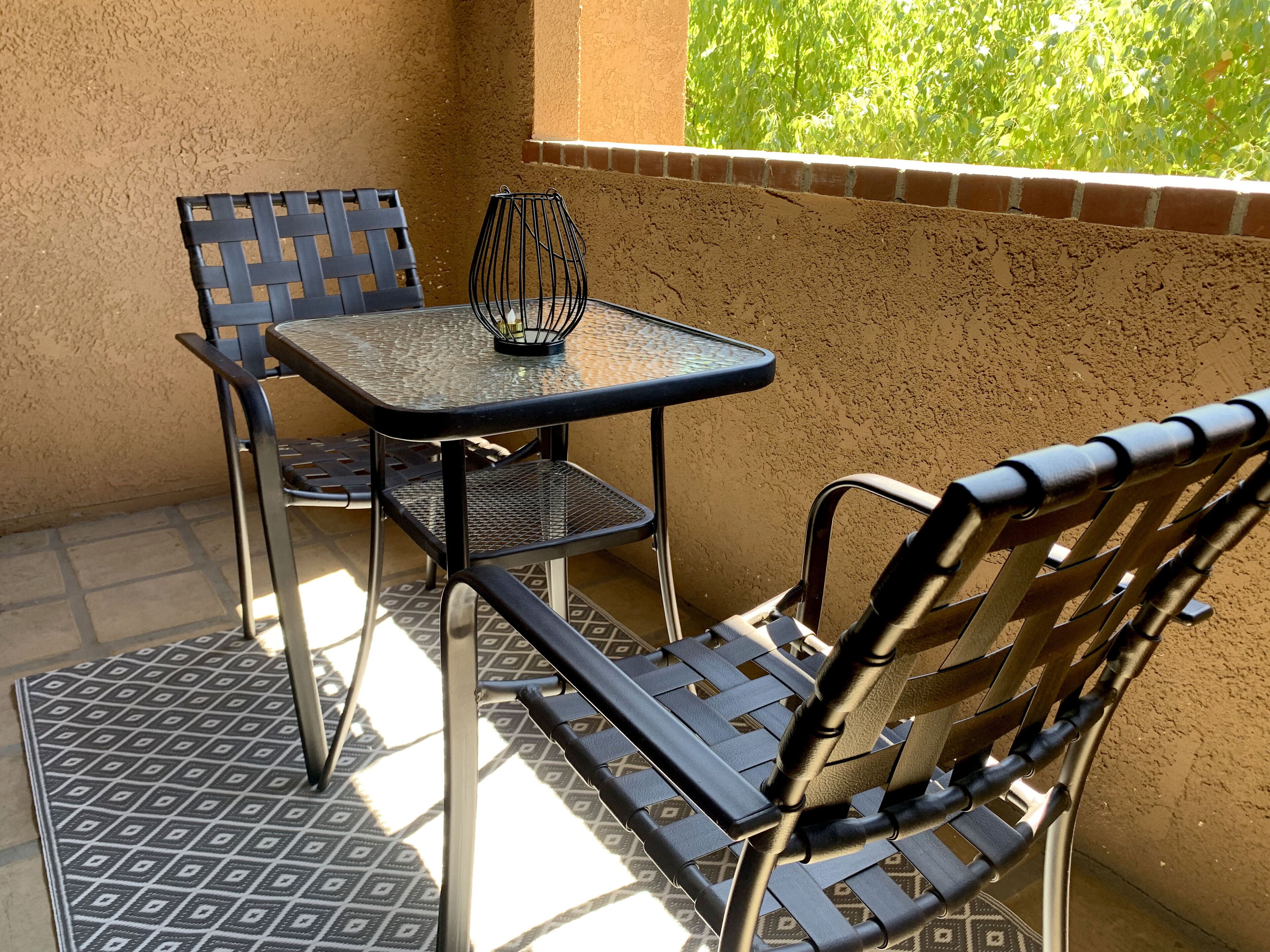 Een klein terras buiten met een tafel en twee stoelen, een decoratieve metalen vaas, en uitzicht op groen door een raam.