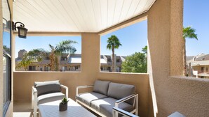 A balcony with a sofa, chairs, and a table, overlooking a residential area with palm trees and houses.