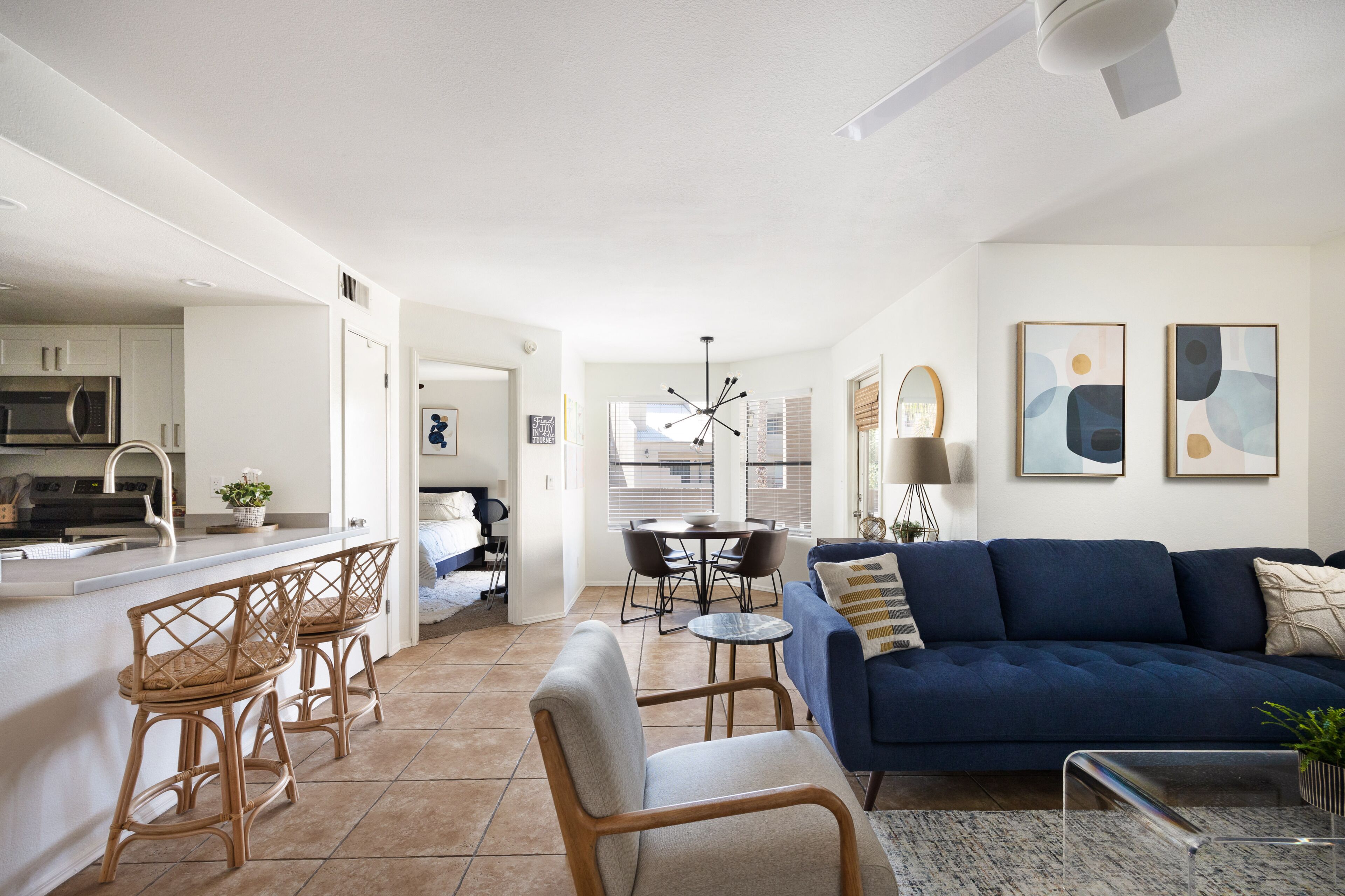 A modern kitchen with white cabinets, a built-in microwave, and a bar area with wicker chairs. There is a dining area with a round table and chairs, and a blue sofa in the living room.