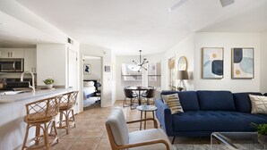 A modern kitchen with white cabinets, a built-in microwave, and a bar area with wicker chairs. There is a dining area with a round table and chairs, and a blue sofa in the living room.