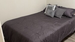 Un lit simple avec une couvre-lit gris foncé et plusieurs oreillers, dans une pièce aux murs de couleur claire et au plancher en bois.