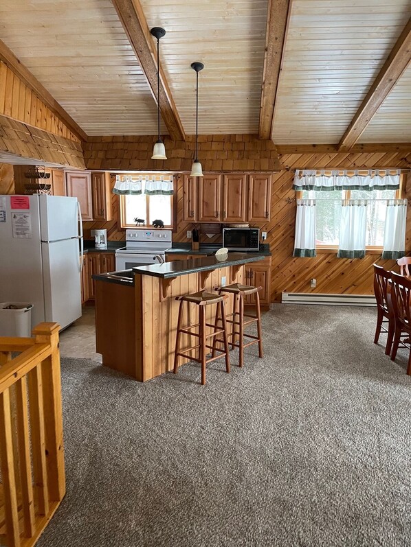 A cozy cabin-style kitchen with wooden walls, a central island with stools, a refrigerator, stove, microwave, and hanging pendant lights.
