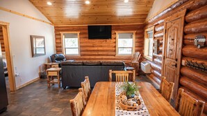 A log cabin interior with a wooden dining table, chairs, a sofa, a television, and a fireplace.