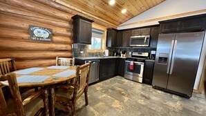 A log cabin-style kitchen with wooden walls, a dining table with chairs, a microwave, a refrigerator, and a stove.