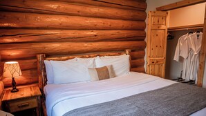 A wooden log cabin-style bedroom with a bed, a nightstand, a lamp, and a wooden wardrobe with clothes hanging inside.