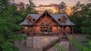A log cabin with a stone foundation, a central chimney, and a porch with railings. - 3 King Suites, Hot Tub, Firepit, Playset, Dogs OK (Broken Bow)