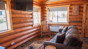 A log cabin interior with a wooden floor, a sofa, a rocking chair, and a window with a view of the outside.
