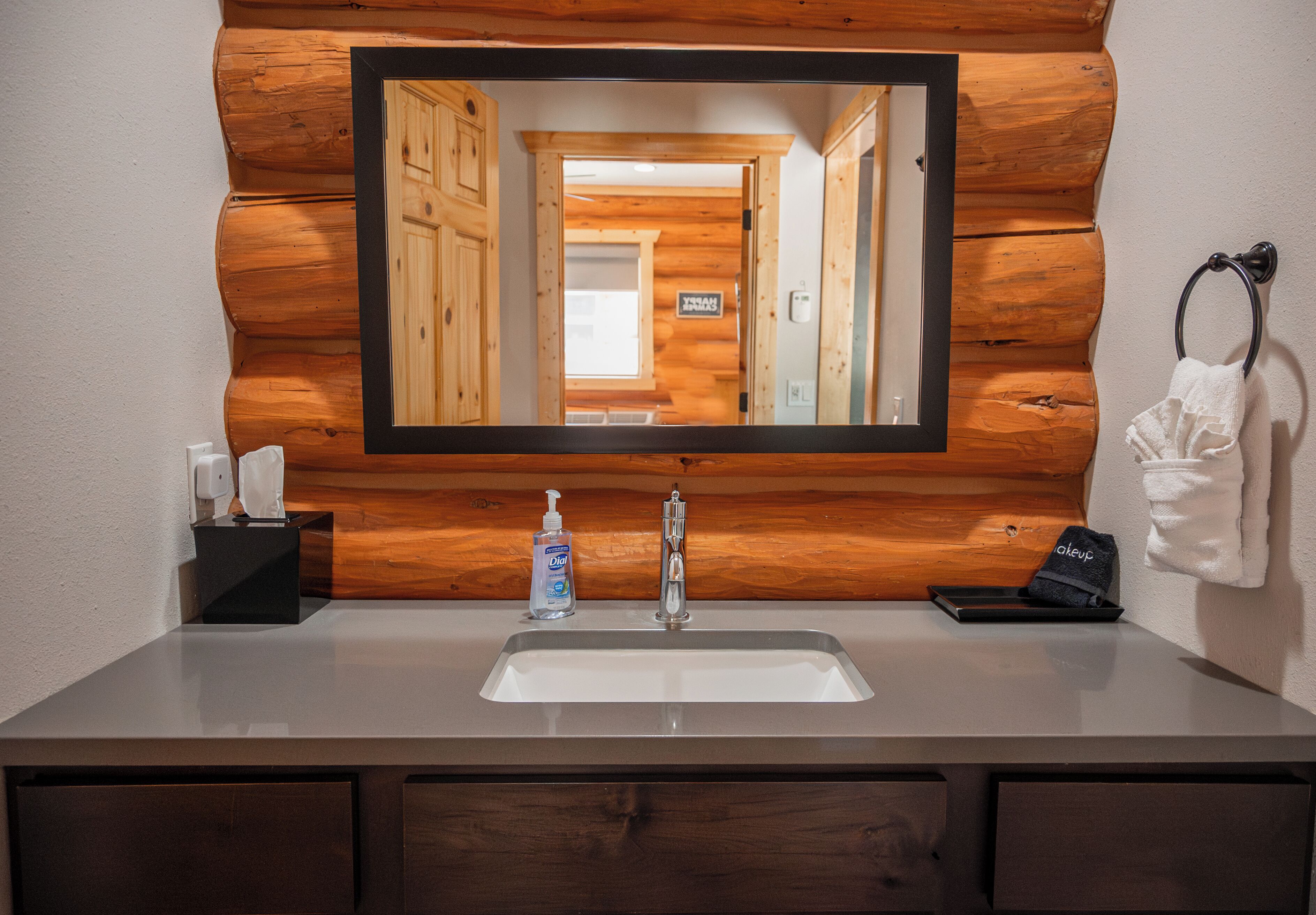 Un baño con pared de tronco de madera, un espejo grande, un lavabo con grifo, un aro para toallas y una botella de jabón de manos.
