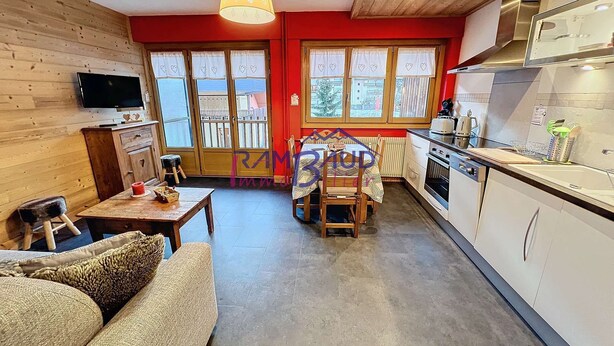 A cozy living room with a sofa, a dining table, and a TV. The kitchen has white cabinets, a stove, and a window with curtains.