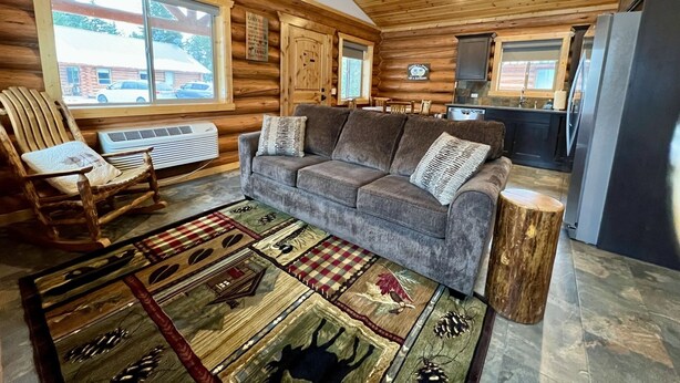 A cozy cabin interior with a wooden ceiling, a gray sofa, a woven rug, and a dining area with wooden chairs.
